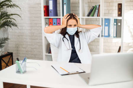 Female doctor wearing protective mask is feeling despair reading bad news online sitting at the office with laptop, a medic holds head with a hands. Virus outbreak conceptの写真素材