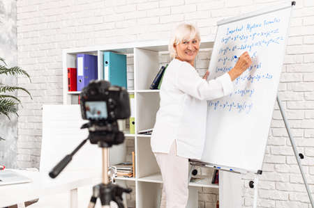 Cheerful senior female teacher writes algebra formulas on a whiteboard in modern classroom and records it on video. Online classes, online studying conceptの写真素材