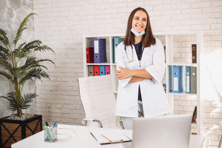 Portrait of young cheerful female doctor wearing medical mask on the chin at the clinic office. A woman physician stands with arms crossed and looks at camera with a friendly smileの写真素材