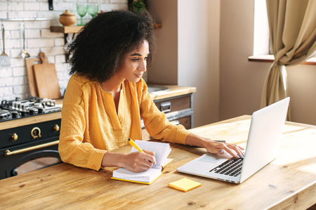 An african-american young woman is using laptop computer for remote work or studying online, she takes notes watching webinars or classes at kitchen table at homeの写真素材