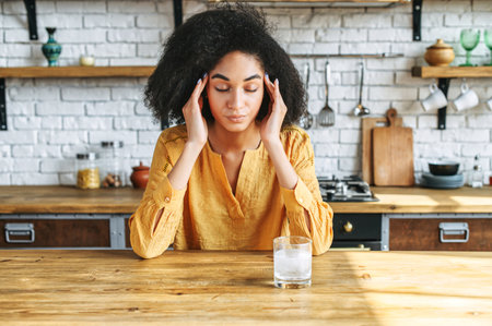 An african american woman suffering from headache and holds temples with hands, a glass of water with effervescent tablet on the table. Drug, remedy from painの写真素材
