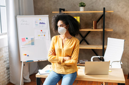 An african american woman wearing protective face mask at the contemporary office. Safeness from viral threat at officeの写真素材