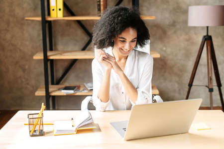 Charming female office employee has online video meeting on laptop. A black smiling woman in smart casual wear talking via video call at modern officeの写真素材