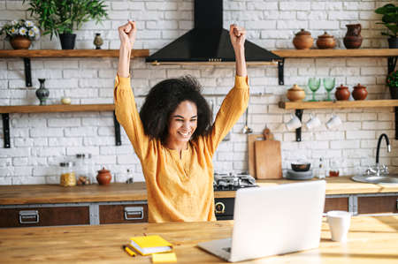 Excited about good news or deal young black woman sits at the desk in home office and looks at laptop screen, holding hands up in triumph gesture. Victory conceptの写真素材