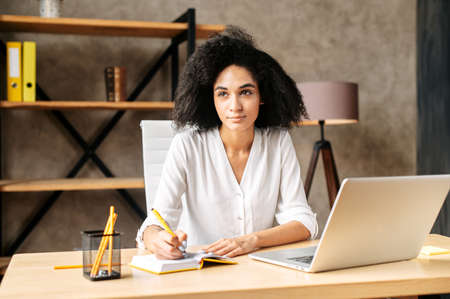 A beautiful black woman in smart casual wear is using laptop computer at a modern office, a thoughtful female employee is writing in plannerの写真素材