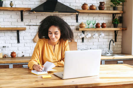 A beautiful black woman in smart casual wear is using laptop computer in the kitchen, a thoughtful female employee is writing in planner. Work remotely from homeの写真素材
