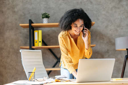 An african american woman is busy at work. A black girl stands near office desk, is talking on phone and taking notes in same times. Attractive young employee takes calls at workの写真素材