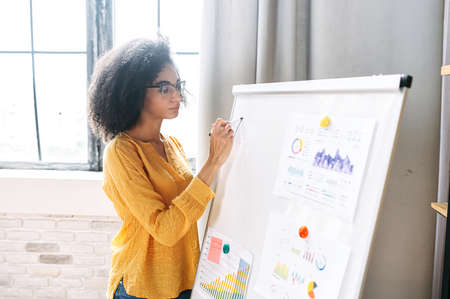 Intelligent african american woman in eyeglasses stands near whiteboard with a graphs and charts on it. Online classes, business trainingの写真素材