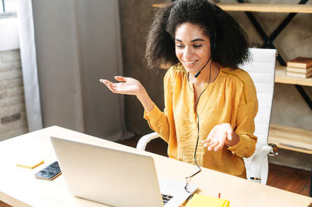 Positive african american woman is using handfree headset for communication sitting at the desk with laptop at office modern space. Call center operator at workの写真素材