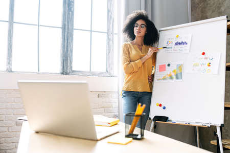 Smart african american woman conducts online webinar, a female coach is explains something on the flip chart in front of laptopの写真素材