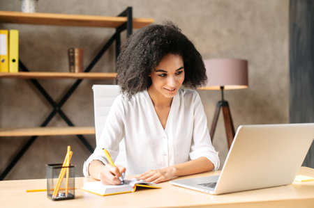 A young african woman looks at laptop screen and takes a note sitting in contemporary officeの写真素材