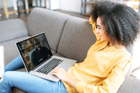 Cheerful African-American girl lays on the comfortable sofa in stylish living room and using trendy laptop for video call to male friend or colleague. Remote work concept, virtual meetingの写真素材