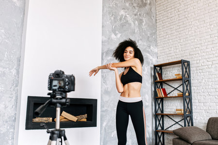 An athletic female fitness coach records fitness video classes at home. An african american girl in sportswear is doing stretching exercises in front of camera. Sports vlogging concept, online trainingの写真素材