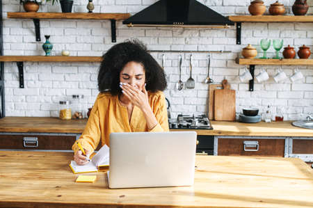 Sleepy an african-american young woman yawns using laptop  for remote work or studying online at homeの写真素材