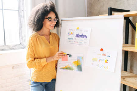 Intelligent african american woman in eyeglasses stands near whiteboard with a graphs and charts on it.の写真素材