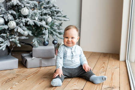 Funny cute baby boy in blue clothes sits indoors on the floor near festive Christmas tree and smilesの写真素材