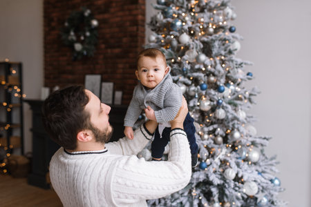 Caring young father is holding little infant son in his arms, a festive Christmas tree on the background. New Years moodの写真素材