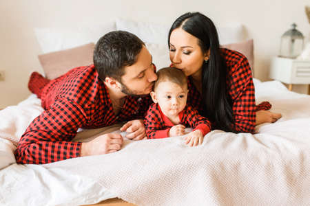 Happy young family of three in red plaid pajamas in bed. A young beautiful mother and a bearded father are kissing cute baby sonの写真素材