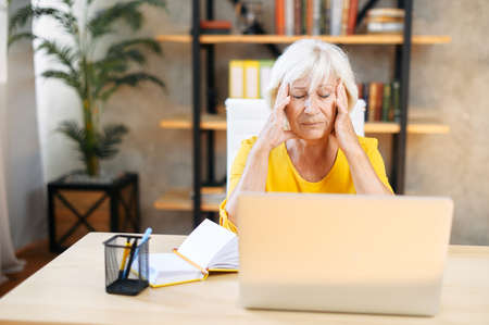 An elderly woman holding head with hands while sitting and working with a laptop indoors. Tired from work exhausted senior woman suffers from headacheの写真素材