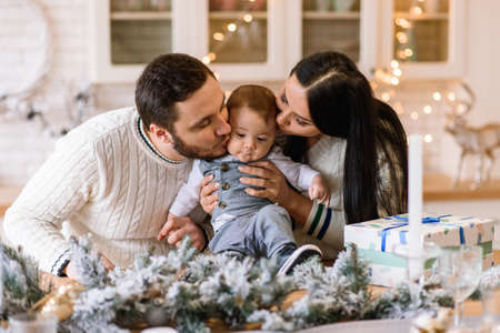 A young parents are kissing little son. Happy family in the kitchen near table decorated with pine branches and candles for Christmasの写真素材