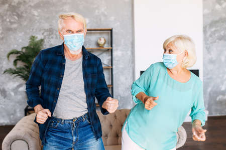 Positive and cheerful senior couple wearing medicine masks has fun together at home during quarantine. Elderly spouses dancing with surgery masks on their facesの写真素材