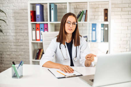 Confident female doctor in eyeglasses sits in the office and using laptop for online consultation. A woman in white formal coat looks at webcam and talking. Virtual visit conceptの写真素材