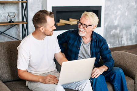 Cheerful two generation men family old senior father and smiling young adult son talking sitting on the comfortable sofa at home, using laptop for websurfing, online shoppingの写真素材