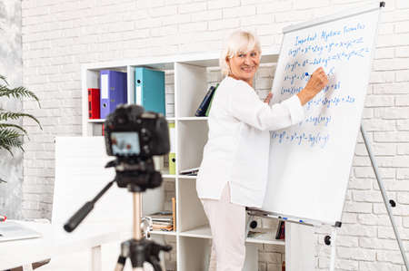 Cheerful senior female teacher writes algebra formulas on a whiteboard in modern classroom and records it on video. Online classes, online studying conceptの写真素材