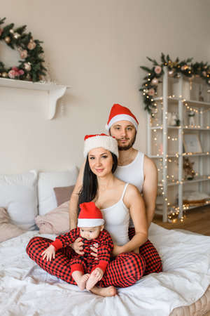 Christmas holidays at home. A young family of three - a father, a mother and cute little son are in santa hats in the bed, a bedroom decorated with Christmas wreath and lights garlandsの写真素材