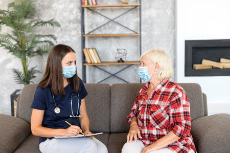 Young serious female doctor in protective medical mask exams senior patient at home appointment and taking a notesの写真素材