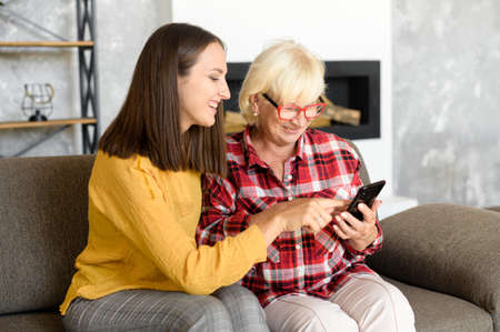 Granddaughter teaching grandmother how to use smartphone. Senior and young woman sit together on...