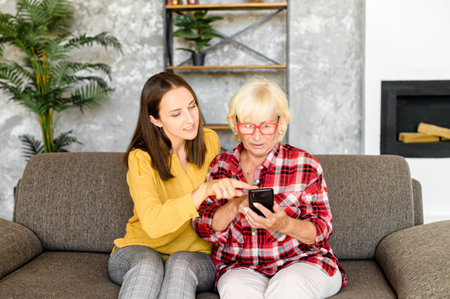 Adult granddaughter teaching the grandmother to use a modern smartphone. Two woman different generations sit on the couch and look at phone screen with interestの写真素材