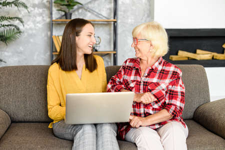 Senior mother and adult daugther sit on the sofa at home and look with interest on computer screen....