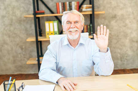 Video call with a mature businessman. A senior grey haired man in smart casual wear looks at webcam and wavingの写真素材