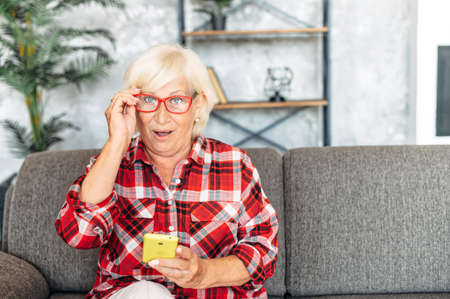 Surprised elderly woman with a phone in her hands sits on the sofa at home. Grandmother saw...