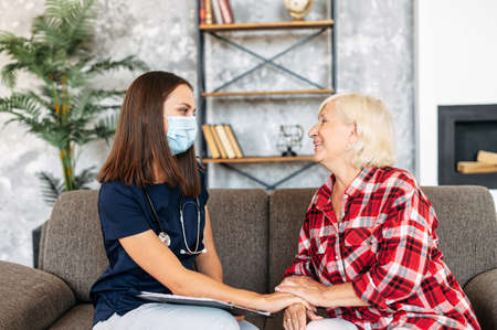 Doctor in protective medical mask visits her senior patient at home. Therapist is taking a notes, a grandmother is complaint on a diseaseの写真素材