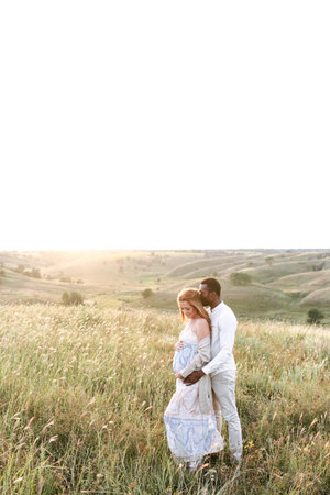 Relaxed multiracial pregnant couple in the park outdoor. An african american husband hugs from the back his charming pregnant wifeの写真素材