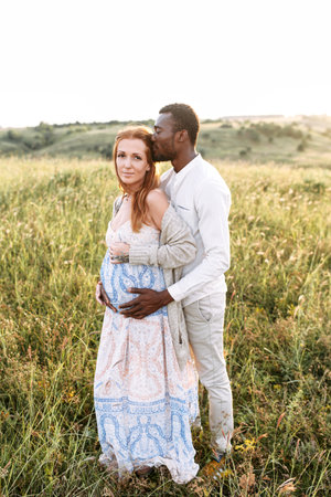 Multiethnic spouses are waiting for a baby. A beautiful pregnant wife and handsome husband in summer wear stand in embrace outdoorsの写真素材
