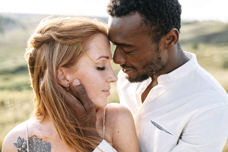 Touching portrait of multi ethnic couple in love outdoor. A handsome black guy is tenderly touching face of charming red haired caucasian woman in nature sunset lightの写真素材