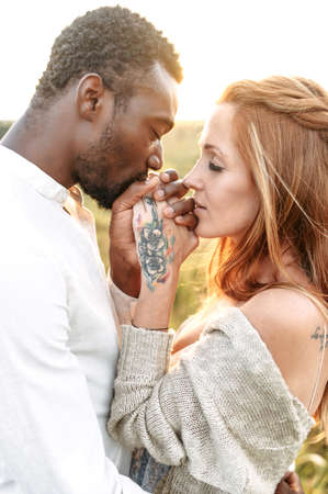 Vertical touching portrait of multiracial couple in sunset light. An african-american man is kissing hands of charming redhaired womanの写真素材