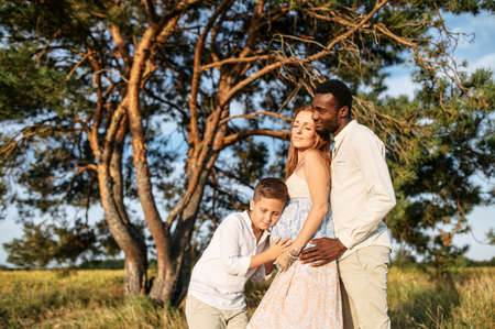 Happy diverse family spends time outdoors together. Pregnant wife, care husband and school age boy on the meadow in warm summer dayの写真素材