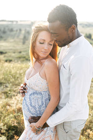 International couple in love walking on the meadow. A white pregnant wife and black husband stand in embrace among nature in warm summer dayの写真素材