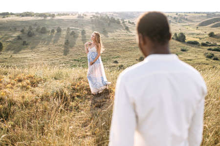 International couple in love walking on the meadow. A white pregnant wife stands on the meadow, a black husband looks at herの写真素材