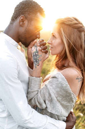 Sun rays shine through a couple in love. An african-american guy is tenderly kissing hands of charming caucasian womanの写真素材