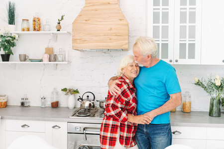 Senior spouses spend time with each other in the kitchen at home. A happy elderly couple in love in embraceの写真素材