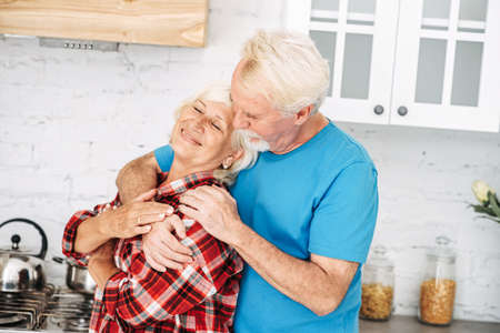 Elderly spouses are hugging in the kitchen. Gray-haired couple in casual plaid shirts spend time together at homeの写真素材