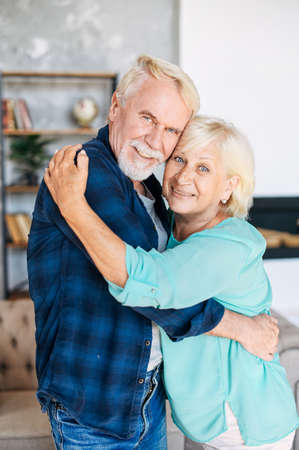 Older couple in love. Cheerful elderly couple smiling and hugging at cozy living roomの写真素材