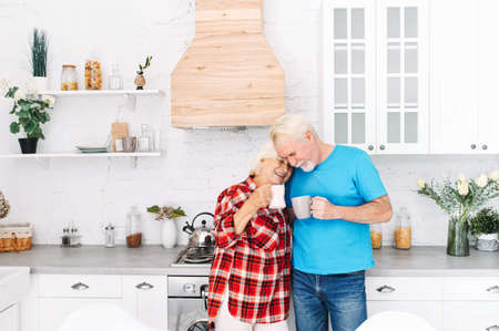 Senior couple is enjoying time together at home. Grandparents with cups of tea in the kitchen are embracingの写真素材