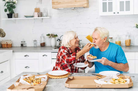 Senior couple in love eating pizza together in the kitchen at home. Beautiful elderly wife is feedin gpiece of pizza her grey-haired mature husband