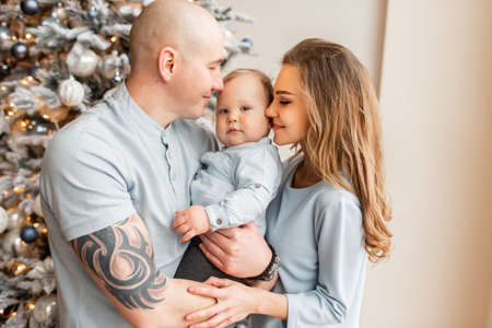 A happy family of three at home near festive Christmas tree. Young parents look at cute baby boy. New Year moodの写真素材